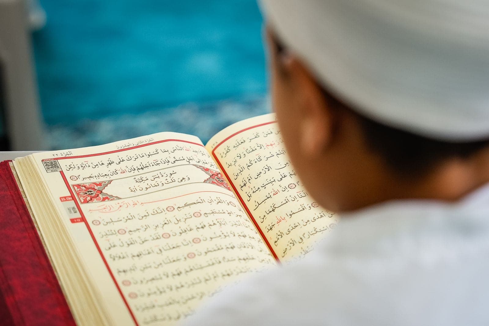 A boy in a white cap reading from an open Quran